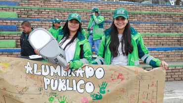 Gestores de alumbrado público sosteniendo cartelera hecha por los niños y niñas y con una luminaria real en las manos.